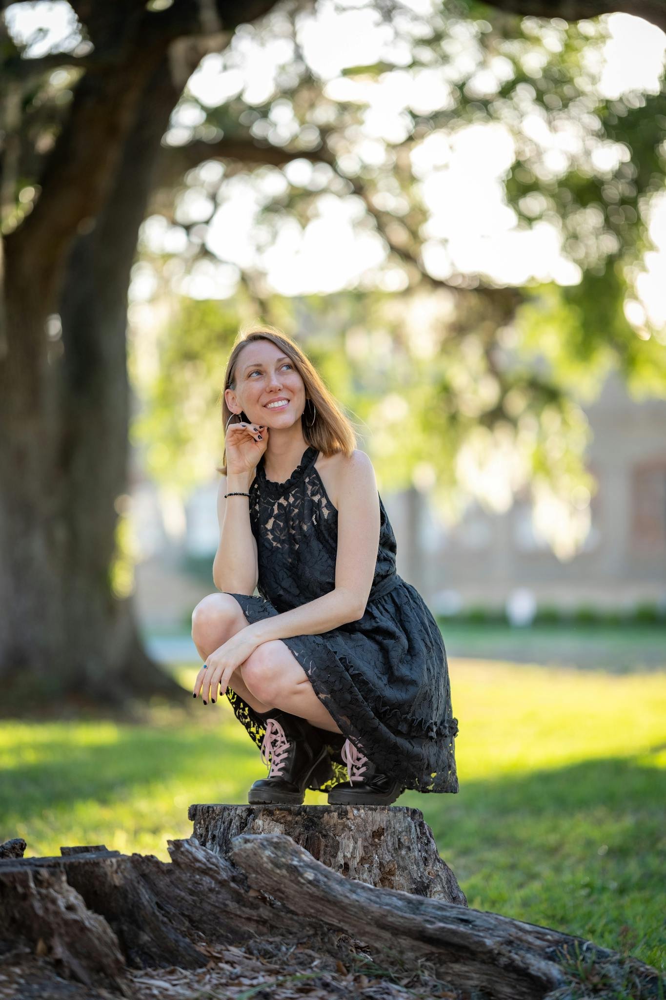 A woman in a black dress smiles while squatting on a tree stump in a sunny park.