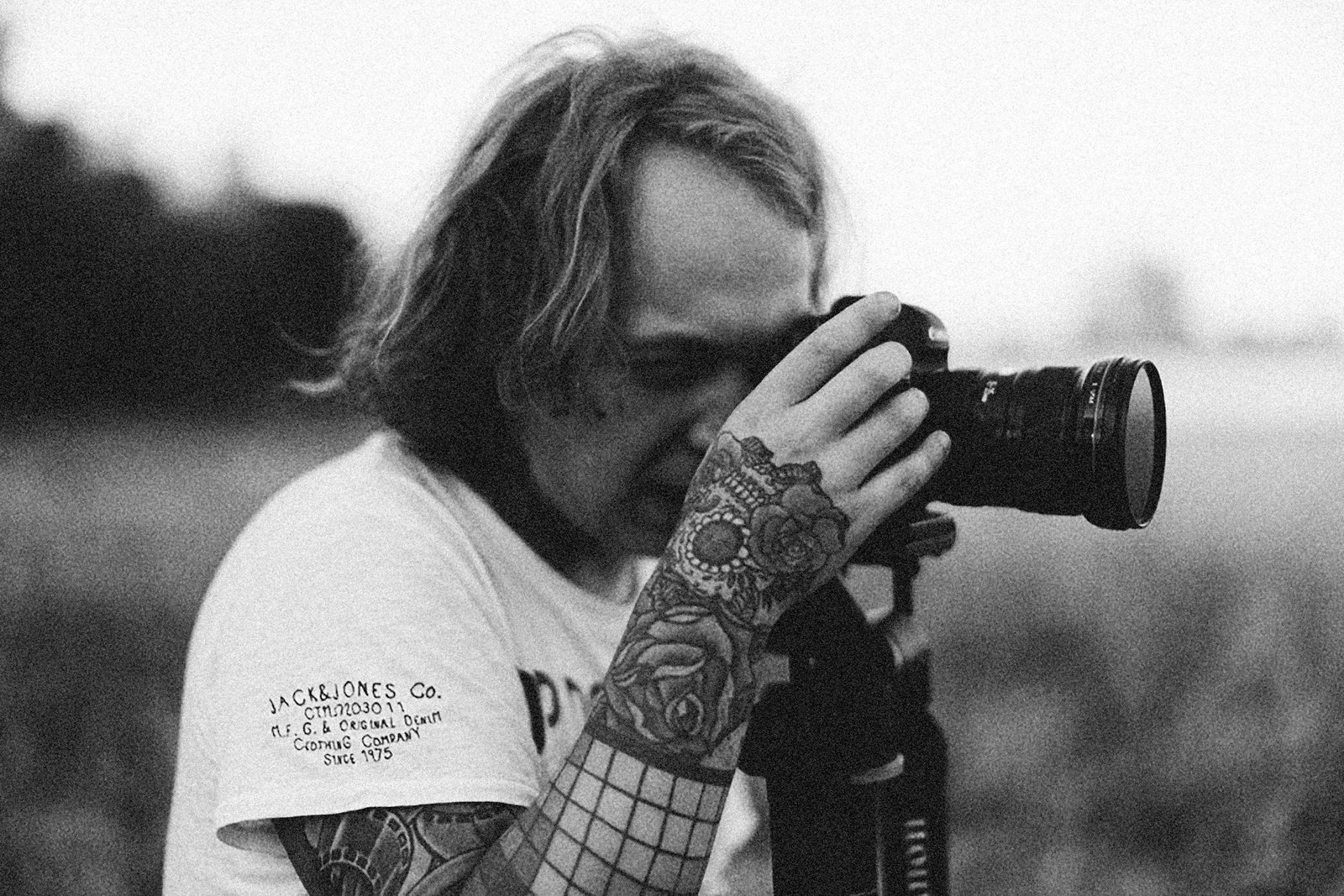Tattooed man takes a monochrome photo outdoors with a tripod camera.